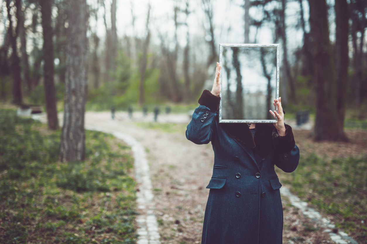 A woman in a green coat standing in a park area holding up a mirror that hides her face